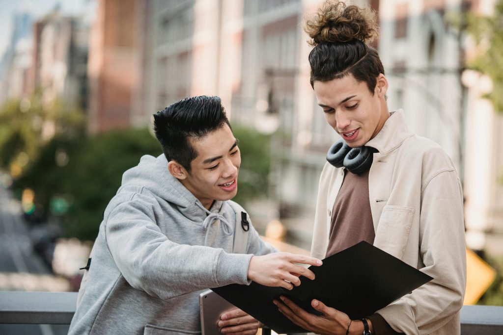 Positive young multiracial male students in casual outfits looking at papers on research in folder while discussing details